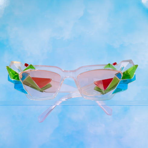 A pair of geometric sunglasses with angular frames featuring vibrant green, blue, and pink accents. The lenses are tinted light pink, adding a playful, modern twist to the bold, colorful design. These sunglasses make a striking, artistic statement